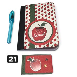May include: A notebook with a red apple pattern, a black spine, and a circular emblem featuring a red apple and the text First Day Of School. Beside it is a translucent blue pen and a small rectangular card with a red apple and two gold stars.