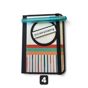 May include: A rectangular notebook with a black elastic band and a translucent blue pen. The cover features a white circle with the text "you are amazing" on a black rectangular label, above horizontal orange and green stripes and vertical lines.