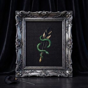 Könnte beinhalten: Gerahmtes Kunstwerk mit einem schwarzen Feder-Motiv mit goldenen Akzenten, umschlungen von einer grünen Schlange. Das Kunstwerk ist vor einem dunklen Hintergrund in einem verzierten Silberrahmen platziert, was eine gotische Ästhetik erzeugt.