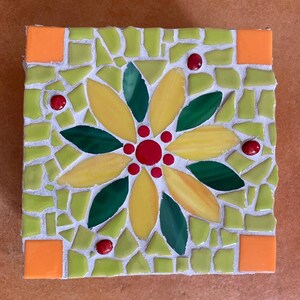 Può includere: Un sottobicchiere a mosaico quadrato con un disegno floreale centrale composto da petali in vetro giallo e verde scuro, circondato da pezzi di vetro verde chiaro. Gli angoli sono accentuati da quadrati arancioni e piccole perle rosse.