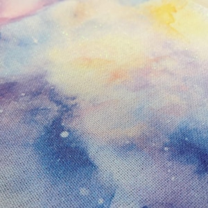 May include: Close-up of a fabric with a watercolour-style design. The pattern features a blend of colours, including blues, purples, pinks, and yellows, creating a dreamy, celestial effect. The fabric has a textured appearance.