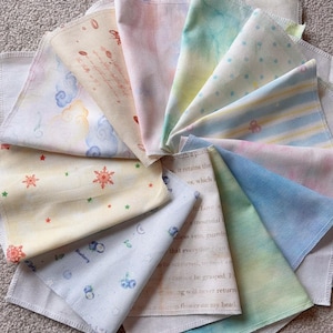 May include: A collection of square fabric cloths arranged in a fan shape, featuring various patterns including clouds, snowflakes, blueberries, stripes, polka dots, and text. The cloths display a range of pastel colors like blue, yellow, pink, and green.