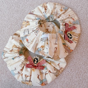 Puede incluir: Dos cubiertas de cuenco de tela color crema con un estampado de Alicia en el país de las maravillas. Cada cubierta tiene un lazo de cinta roja con un adorno negro y dorado. Las cubiertas están recogidas con elástico.