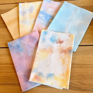 Op de afbeelding: Zes rechthoekige stukken stof met aquarelontwerpen in pasteltinten van geel, roze, blauw en paars. De stoffen zijn gerangschikt op een houten oppervlak. Elk stuk heeft een uniek wolkenachtig patroon, wat een zachte, dromerige esthetiek creëert.