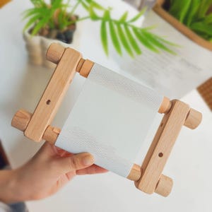 May include: A wooden embroidery frame with a light grey fabric stretched across it. The frame is held in a hand, and the wood has a natural finish. The fabric is a fine weave, suitable for cross-stitch or embroidery projects.