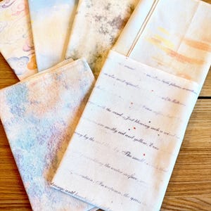 May include: A collection of folded fabric napkins with various watercolor designs. The napkins feature pastel colors, including blue, pink, and yellow, with some displaying text. The napkins are arranged on a wooden surface.