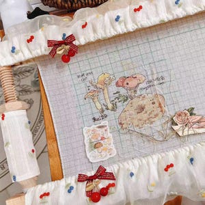May include: A cross-stitch frame with white fabric adorned with embroidered cherries and blue berries. A cross-stitch project with mushroom illustrations is in progress. Decorative red bows with cherry accents add a whimsical touch.