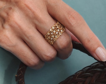 May include: A wide gold band ring featuring an intricate filigree pattern with small, clear, round-cut stones embedded throughout the metalwork. The ring has a textured, scalloped edge design and a polished finish, worn on a finger.