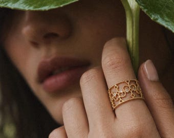 May include: A wide gold band ring featuring an intricate openwork filigree pattern with small circular cutouts and tiny embedded clear stones. The metal edges of the ring are finished with a delicate beaded texture along the top and bottom borders.
