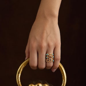 May include: A gold-toned multi-band ring featuring a series of interconnected thin wires adorned with small, round, bezel-set turquoise and dark red garnet gemstones arranged in a repeating pattern across the width of the band.