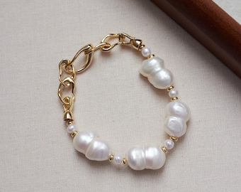 May include: A gold-toned chain bracelet featuring irregular white baroque pearls interspersed with small round white pearls and gold-toned metal beads. The bracelet includes a large gold-toned lobster clasp and a series of textured, organic-shaped links.