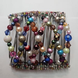 May include: A beaded hair comb with a silver metal base. The comb features a variety of colorful beads in shades of blue, pink, green, and gold. The beads are strung together in a decorative pattern.