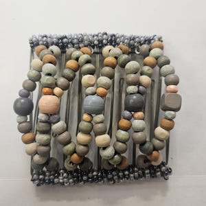 May include: A hair comb with metal tines and decorative wooden beads in various colors, including gray, brown, green, and pink. The beads are strung together in curved lines across the comb.