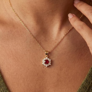 May include: A gold chain necklace featuring a pendant with a central round red gemstone surrounded by a halo of small clear white stones. The pendant is suspended from a delicate gold link chain that rests against the collarbone area.