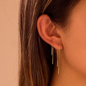 Könnte beinhalten: Goldene Fadenohrringe mit minimalistischem Design. Die Ohrringe haben eine zarte Kette, die vom Ohr herabhängt, mit einem geraden Stab am Ende. Die Ohrringe sind an einem Ohr abgebildet.