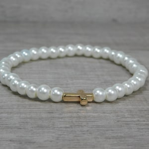 Puede incluir: Pulsera de cuentas blancas con un dije de cruz dorada. La pulsera está hecha de cuentas redondas blancas y tiene un diseño elástico. La cruz dorada está centrada en la pulsera.