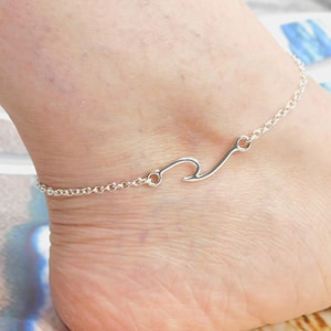 May include: A silver-colored anklet featuring a wave design. The delicate chain wraps around the ankle, with the wave charm positioned on the top of the foot. This piece of jewelry is a stylish accessory.