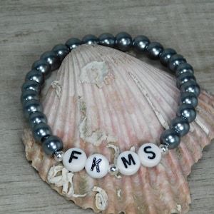 May include: A dark gray beaded bracelet with white letter beads spelling out "FKMS". The bracelet is resting on a seashell. The beads are a dark gray color and the letters are black.