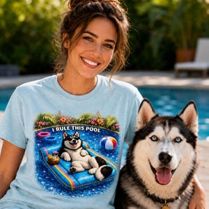 May include: Pale blue t-shirt featuring the text "I RULE THIS POOL" and a graphic of a husky dog on a pool float. The dog wears sunglasses and has a cocktail. A real husky dog with blue eyes is next to the person wearing the shirt.