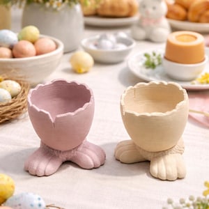 Pode incluir: Dois porta-ovos de cerâmica, um rosa claro e outro creme, cada um em forma de casca de ovo rachada apoiada em patas de coelho. O fundo inclui decorações de Páscoa, como ovos, flores e uma figura de coelho.