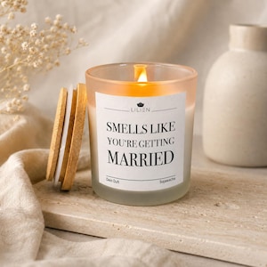 Könnte beinhalten: Eine brennende Kerze in einem klaren Glasgefäß mit einem weißen Etikett, das den Text "SMELLS LIKE YOU'RE GETTING MARRIED" trägt, sowie einem daneben liegenden Holzdeckel. Die Kerze besteht aus Sojawachs und zeigt den Markennamen LILIEN auf dem Etikett.