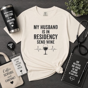 Könnte beinhalten: Ein cremefarbenes T-Shirt mit schwarzem Text MY HUSBAND IS IN RESIDENCY SEND WINE über einem Weinglas und einer Herzschlaglinie. Zubehör umfasst einen schwarzen Becher, einen Ausweishalter, eine Tragetasche, ein Notizbuch und ein Stethoskop.