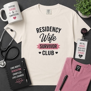 Könnte beinhalten: Ein cremefarbenes T-Shirt mit dem schwarzen und rosa Text "RESIDENCY Wife SURVIVOR CLUB". Das Zubehör umfasst einen weißen Becher mit "CAFFEINE SCRUBS & COMPASSION", ein schwarzes Stethoskop, ein schwarzes Notizbuch, ein rosa Kasack-Oberteil und einen Ausweishalter.