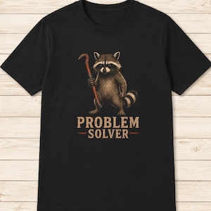 May include: A black short-sleeved t-shirt featuring a graphic illustration of a raccoon holding a red crowbar. Below the raccoon, the text "PROBLEM SOLVER" is printed in a distressed, light-colored font with horizontal lines on both sides.
