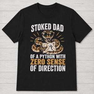 May include: A black short-sleeve t-shirt featuring a graphic of a brown python wearing sunglasses and holding a map with a question mark. The text reads STOKED DAD OF A PYTHON WITH ZERO SENSE OF DIRECTION in white and yellow block lettering.