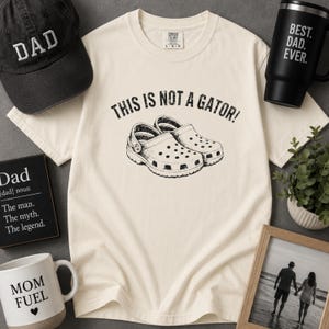 Könnte beinhalten: Ein cremefarbenes Kurzarm-T-Shirt mit einer schwarzen Grafik von zwei Clogs und dem Text THIS IS NOT A GATOR! darüber. Das T-Shirt ist umgeben von einer schwarzen Baseballkappe mit DAD-Schriftzug, einem schwarzen Becher mit BEST. DAD. EVER.-Text, einer Tasse mit MOM FUEL-Aufschrift und einem gerahmten Foto.