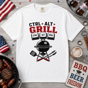 May include: A white short-sleeve t-shirt featuring a graphic print with the text CTRL + ALT + GRILL above three keyboard key icons labeled CTRL, ALT, and GRILL. Below the keys is a black charcoal grill with a spatula and tongs crossed underneath.