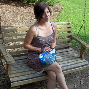 May include: A woman sits on a wooden swing holding a blue and white tote bag with a colorful elephant design. The bag has a black strap and a zipper closure.