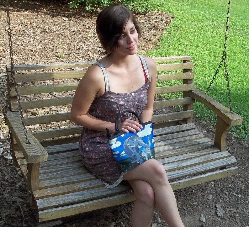 May include: A woman sits on a wooden swing holding a blue and white tote bag with a colorful elephant design. The bag has a black strap and a zipper closure.