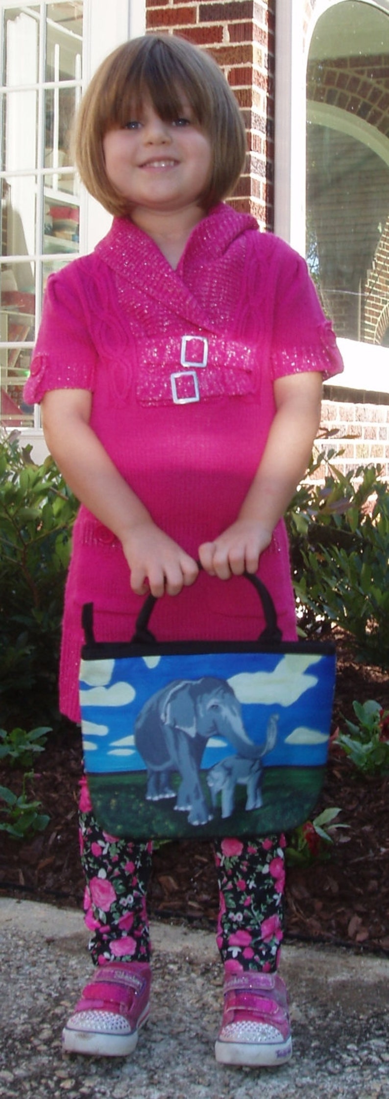 May include: A young girl wearing a pink sweater with a sparkly design and black and white floral leggings is holding a blue and white tote bag with a painted image of an elephant and a baby elephant.