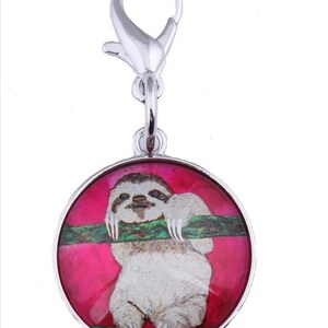 May include: A silver bag charm with a round, pink and green glass charm featuring a sloth hanging from a branch. The charm has a silver clasp.