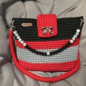 May include: A handmade crochet handbag in black, red, and grey. The bag features a red flap with a silver bow clasp, a beaded strap, and a matching red handle. The bag is a stylish accessory.