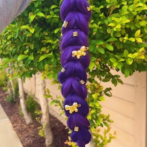 May include: A long, braided purple hair extension adorned with gold-colored hair accessories. The braid is thick and features several decorative gold clips and charms. The hair extension is held against a backdrop of green foliage.
