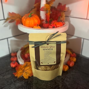 Może przedstawiać: Torba z papieru pakowego z napisem "Autumn Glow Simmer" z wymienionymi składnikami, zawiązana zieloną wstążką. Torba otoczona jest pomarańczowymi dyniami, jesiennymi liśćmi i białą, wielopoziomową tacą z większą ilością dyń i liści.