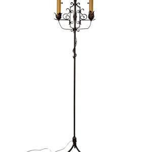 Op de afbeelding: Een hoge vloerlamp van donker metaal met een slanke centrale stang en een decoratief gedraaid rankmotief. Het bovenste gedeelte bevat twee kaarsvormige lampen met oranje glas, ondersteund door sierlijk krulwerk en bladvormige metalen houders.