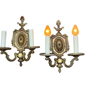 May include: A pair of two-light wall sconces featuring an ornate gold-toned metal backplate with a central oval medallion. Each sconce has two white candle-style sleeves, with one sconce illuminated by two warm orange-toned flame-shaped light bulbs.