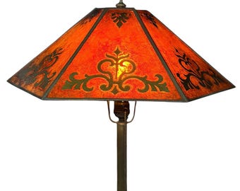 May include: A table lamp with a hexagonal shade featuring a mottled orange surface and dark green decorative scrollwork patterns. The lamp has a slender metal stem and a circular base with a textured, scalloped edge design in a metallic finish.