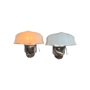 May include: Two wall sconces featuring scalloped, translucent glass shades and polished chrome bases. One shade emits a warm, amber-toned light, while the other is unlit and white. Each fixture includes a small pull chain for operation.
