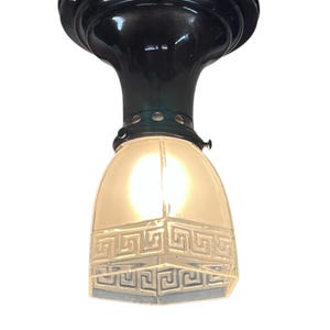 May include: An antique ceiling light fixture with a dark bronze finish. The fixture features a decorative glass shade with a geometric pattern. The light is turned on, illuminating the glass shade.