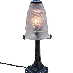 May include: A small, antique-style table lamp with a frosted glass shade featuring a carved floral design. The lamp has a black metal base and stem with a leaf motif. The lamp is lit, casting a warm glow.