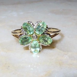 Pode incluir: Um anel de ouro com um design floral com cinco pedras preciosas ovais verdes claras. O centro da flor tem um pequeno diamante redondo. O anel está sobre um fundo branco e dourado.