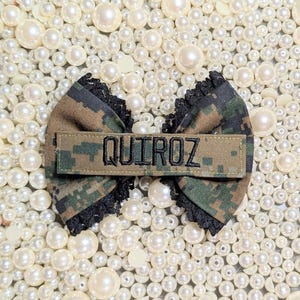 May include: A camouflage bow with black lace trim and a rectangular patch that reads "QUIROZ" in black letters. The bow is set against a backdrop of white pearls.