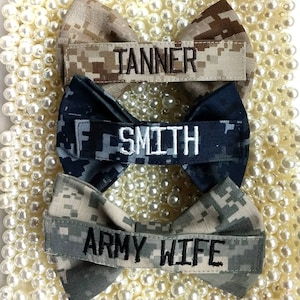 May include: Three bow ties with name patches. The top bow tie is tan and brown camouflage with the name "TANNER". The middle bow tie is navy blue digital camouflage with the name "SMITH". The bottom bow tie is gray and green camouflage with the text "ARMY WIFE".