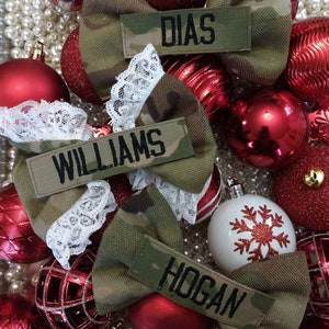 May include: Camouflage fabric bows with white lace and name patches reading "DIAS", "WILLIAMS", and "HOGAN". The bows are set amongst red and gold Christmas baubles and white pearls.