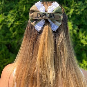 May include: A camouflage-patterned hair bow with white lace trim and the name "CARMONA" embroidered on a rectangular patch. The bow is set against a backdrop of long, light brown hair.