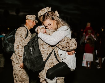 Könnte beinhalten: Eine Frau in einem weißen Pullover umarmt eine Person in Militäruniform. Die Uniform hat ein Tarnmuster. Andere Personen in ähnlichen Uniformen und mit Rucksäcken sind im Hintergrund. Die Szene ist schwach beleuchtet.